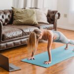 Side view full body of sportive female in activewear practicing kumbhakasana asana on mat near laptop during online lesson in living room near couch