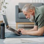 Elderly man practicing yoga on a mat while watching online instructions on a laptop.